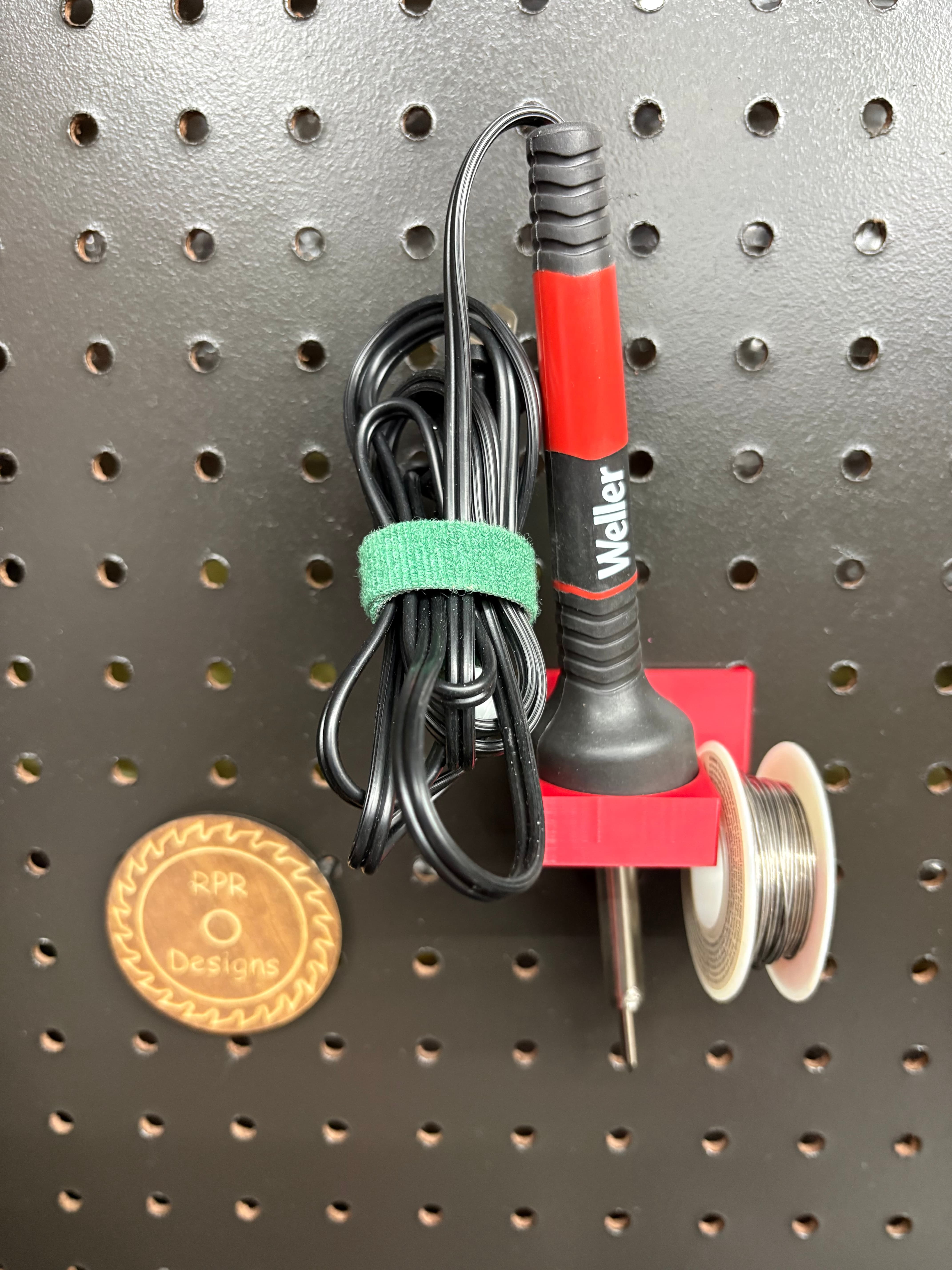 Red 3D printed soldering iron holder with solder spool arm mounted on a pegboard
